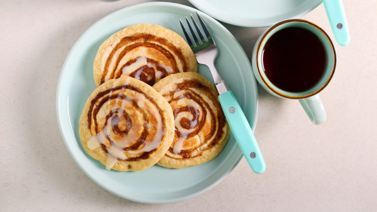 Zimtschnecken-Pancakes auf Teller neben einer Kaffeetasse.