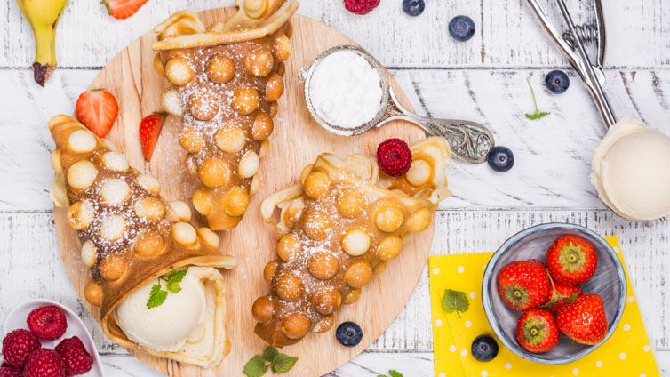 Bubble-Waffeln mit einer Eiskugel gefüllt liegen auf einem Holzbrett, daneben Puderzucker und verschiedene Beeren.