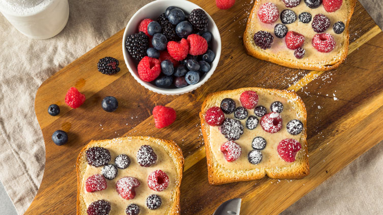 Drei Custard-Toasts mit Beeren und Puderzucker liegen auf einem Holzbrett.