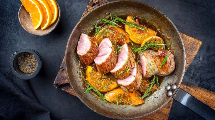 Aufgeschnitte Schweinelende in Orangensauce wird in einer Pfanne auf einem Holzbrett serviert.